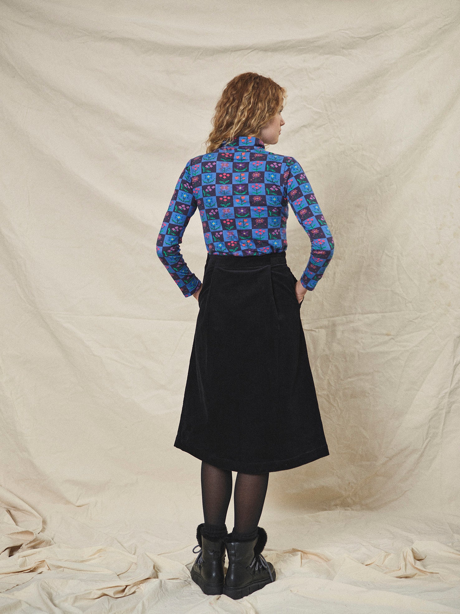 A person wearing a straight black corduroy skirt with a shaped waistband and deep inverted pleats, paired with a multicolored floral top and black boots.