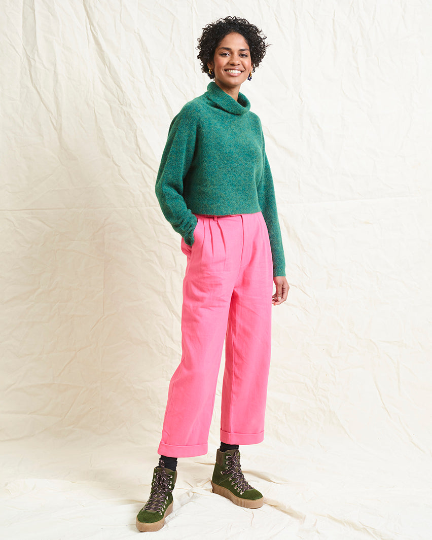 A model wearing bright pink wide leg trousers with pleats and a tab front opening, paired with a green sweater and brown boots.