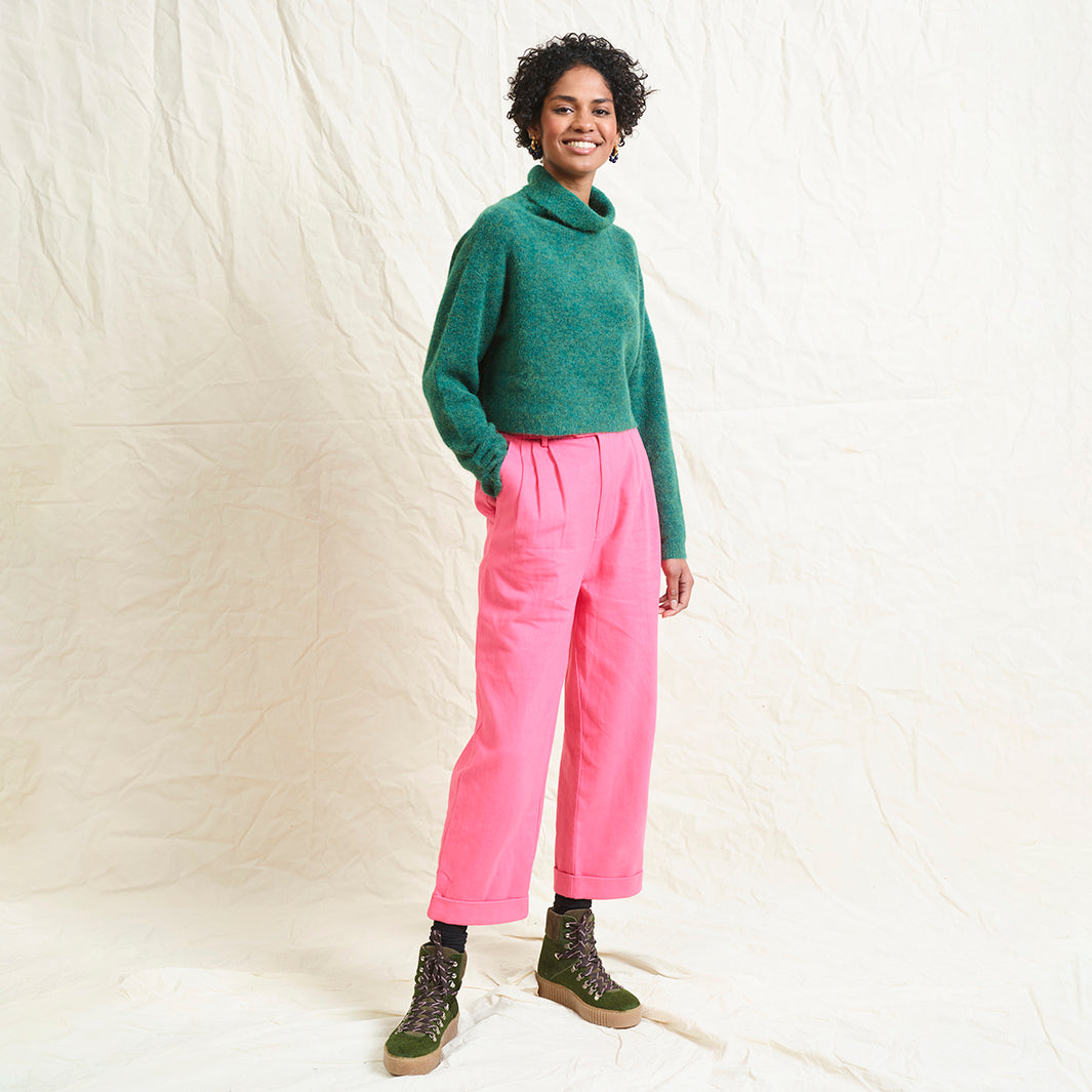 A model wearing bright pink wide leg trousers with pleats and a tab front opening, paired with a green sweater and brown boots.