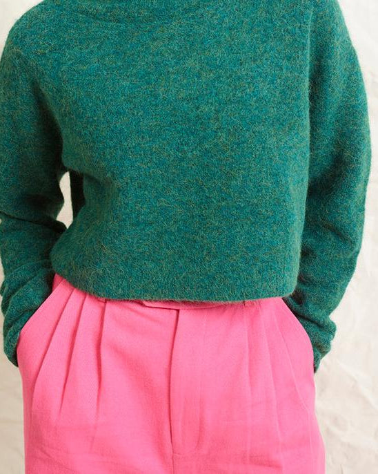 A model wearing bright pink wide leg trousers with pleats and a tab front opening, paired with a green sweater and brown boots.