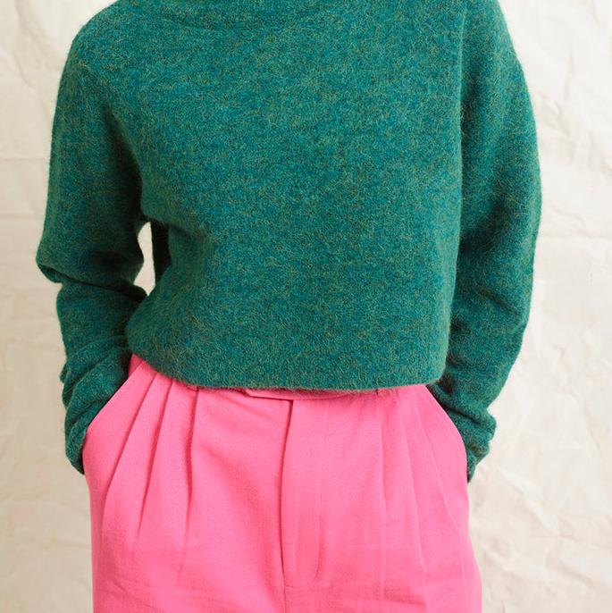 A model wearing bright pink wide leg trousers with pleats and a tab front opening, paired with a green sweater and brown boots.