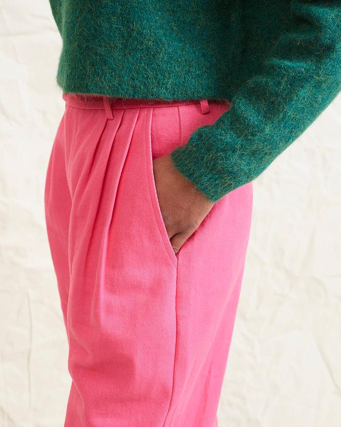 A model wearing bright pink wide leg trousers with pleats and a tab front opening, paired with a green sweater and brown boots.