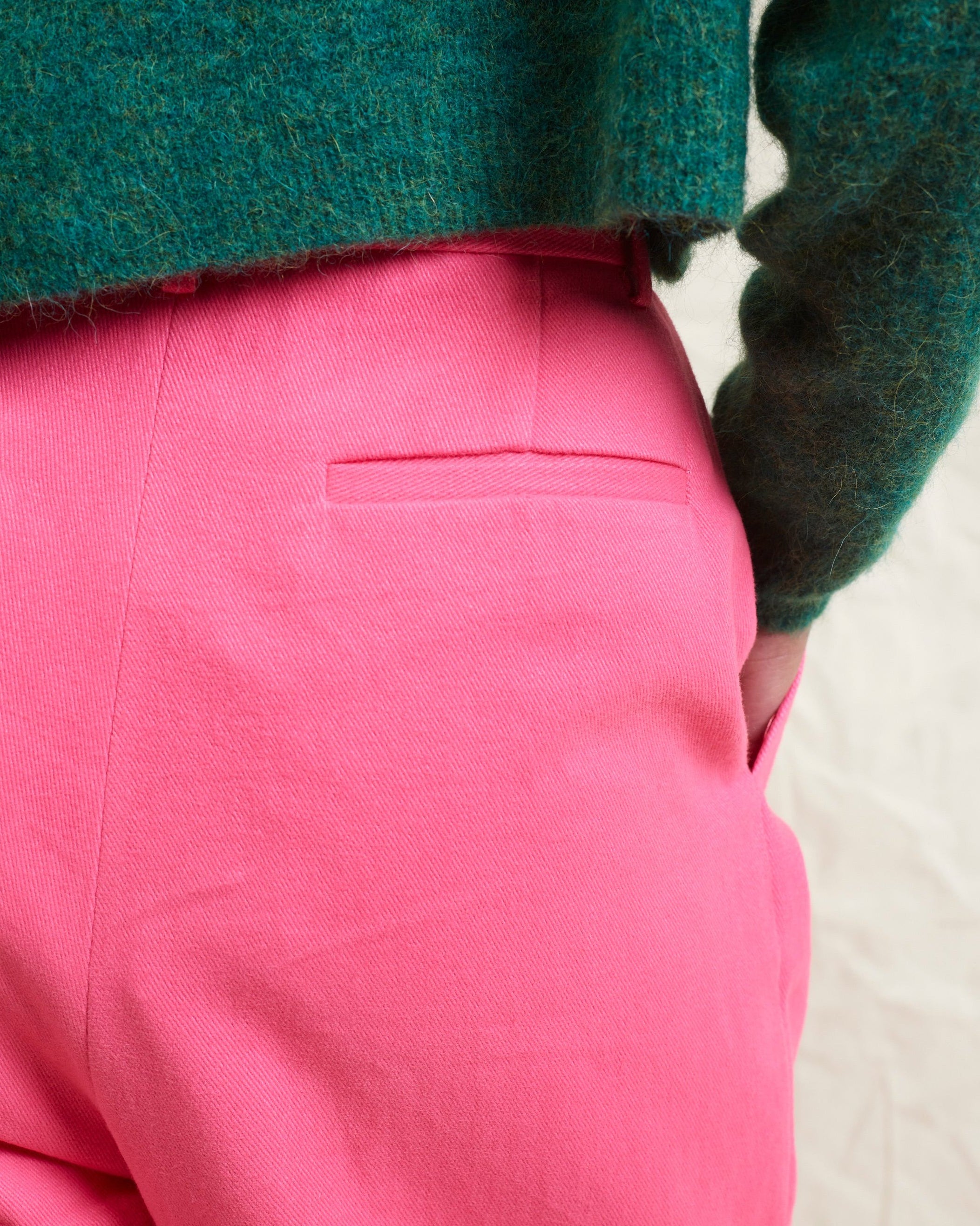 A model wearing bright pink wide leg trousers with pleats and a tab front opening, paired with a green sweater and brown boots.