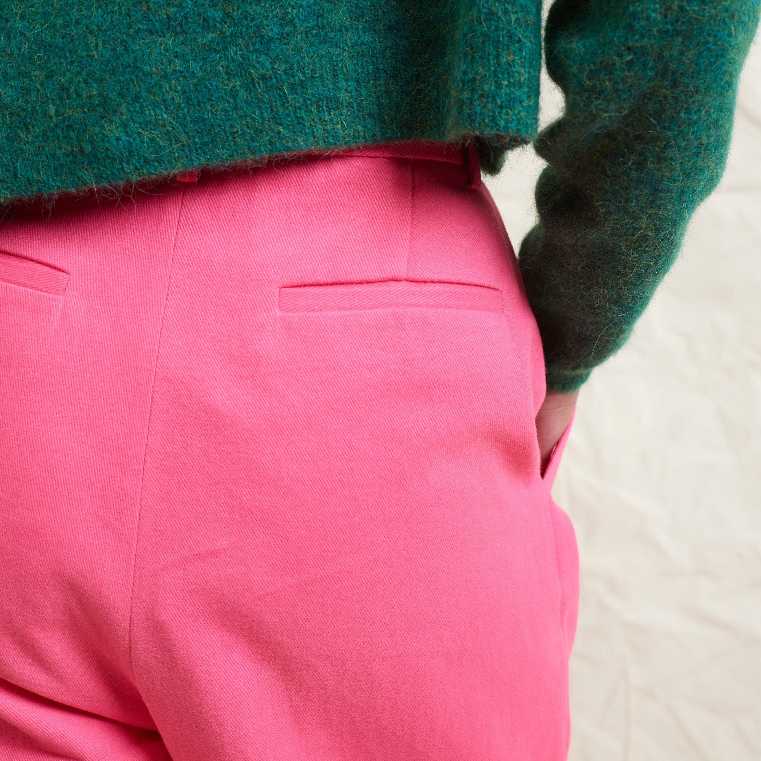 A model wearing bright pink wide leg trousers with pleats and a tab front opening, paired with a green sweater and brown boots.