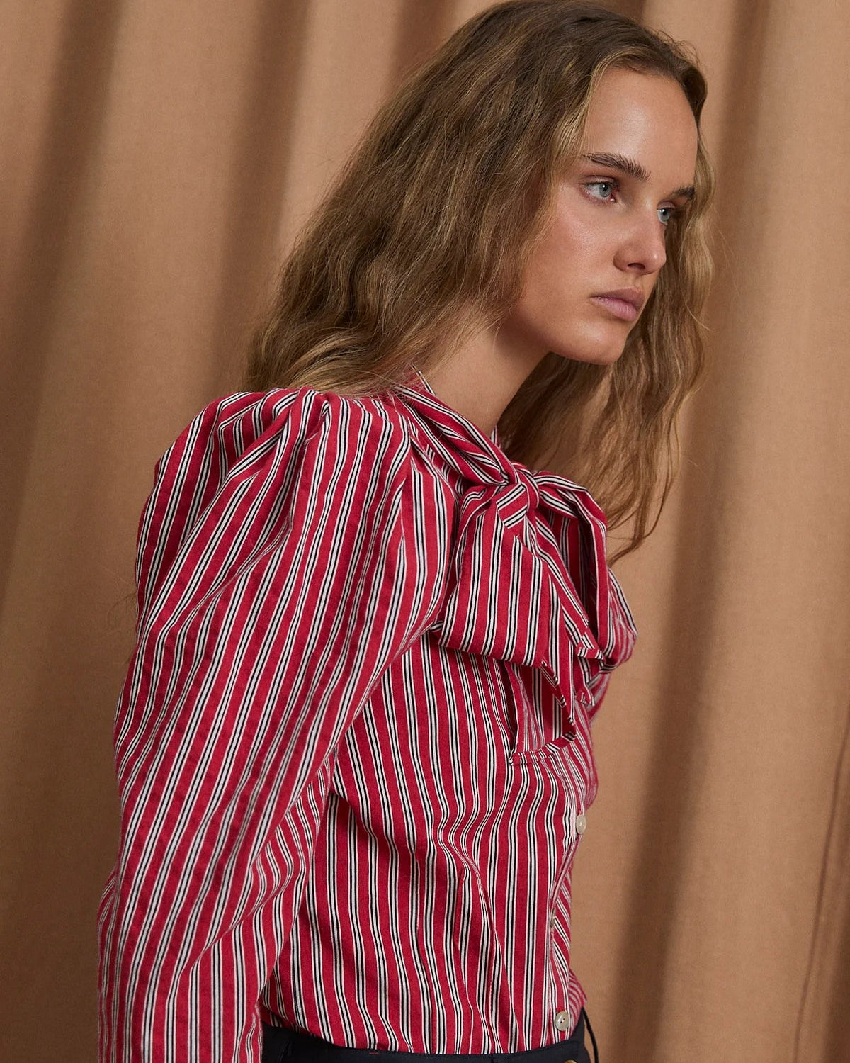 A woman wearing a red and white vertically striped blouse with a large ruffled bow at the collar, paired with dark trousers, posed against a draped tan fabric backdrop.
