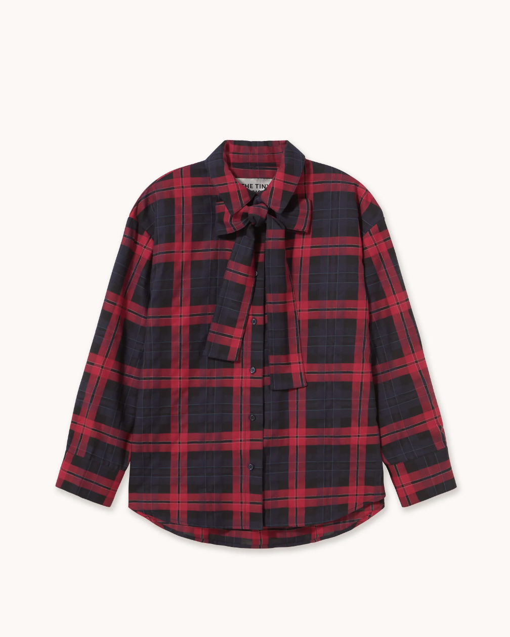 A long-sleeve button-up shirt with a red and black plaid pattern, featuring a self-tie neck bow and a small chest pocket, displayed flat against a light cream background.