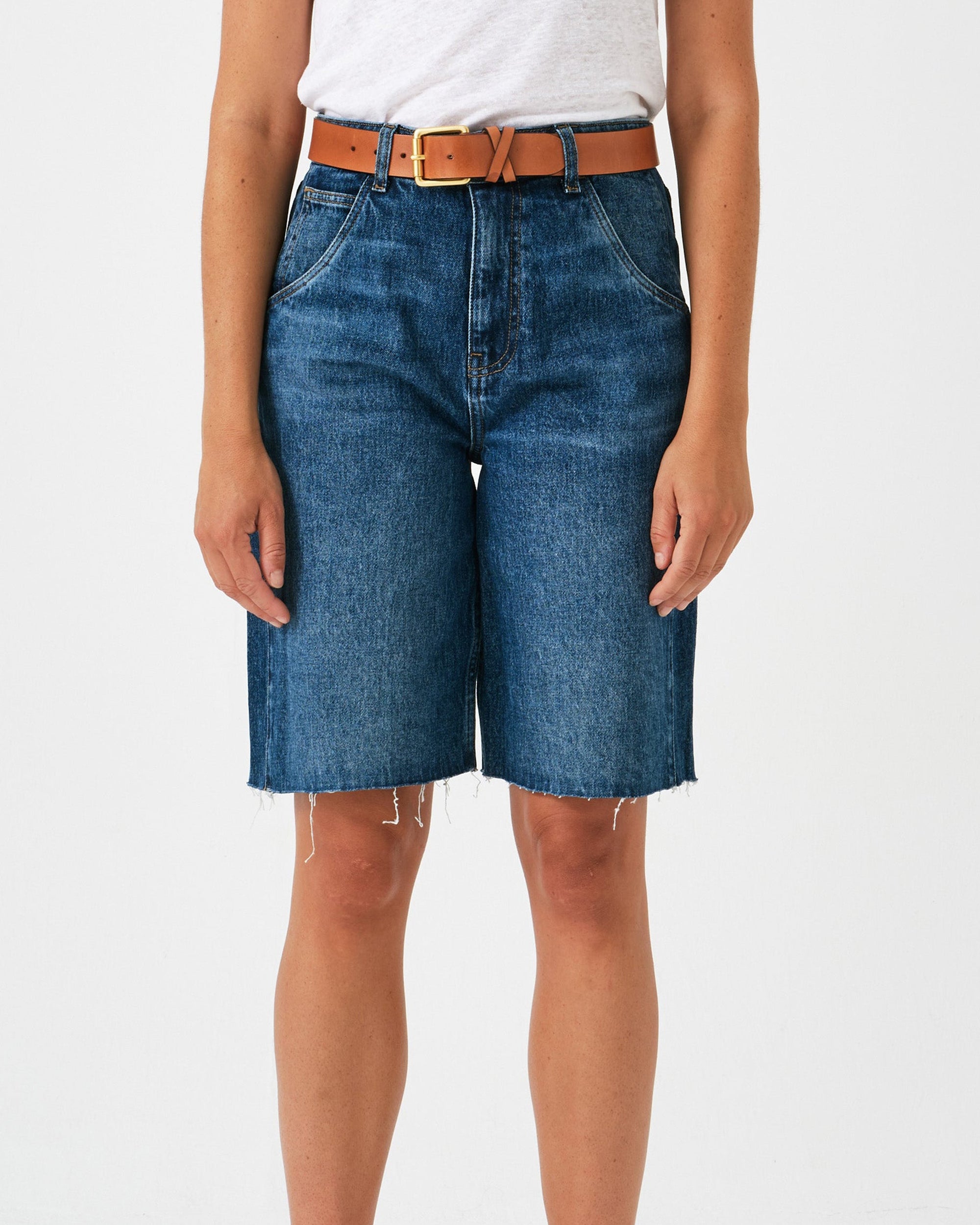 Person wearing blue denim shorts with a white shirt and brown belt on a light gray background