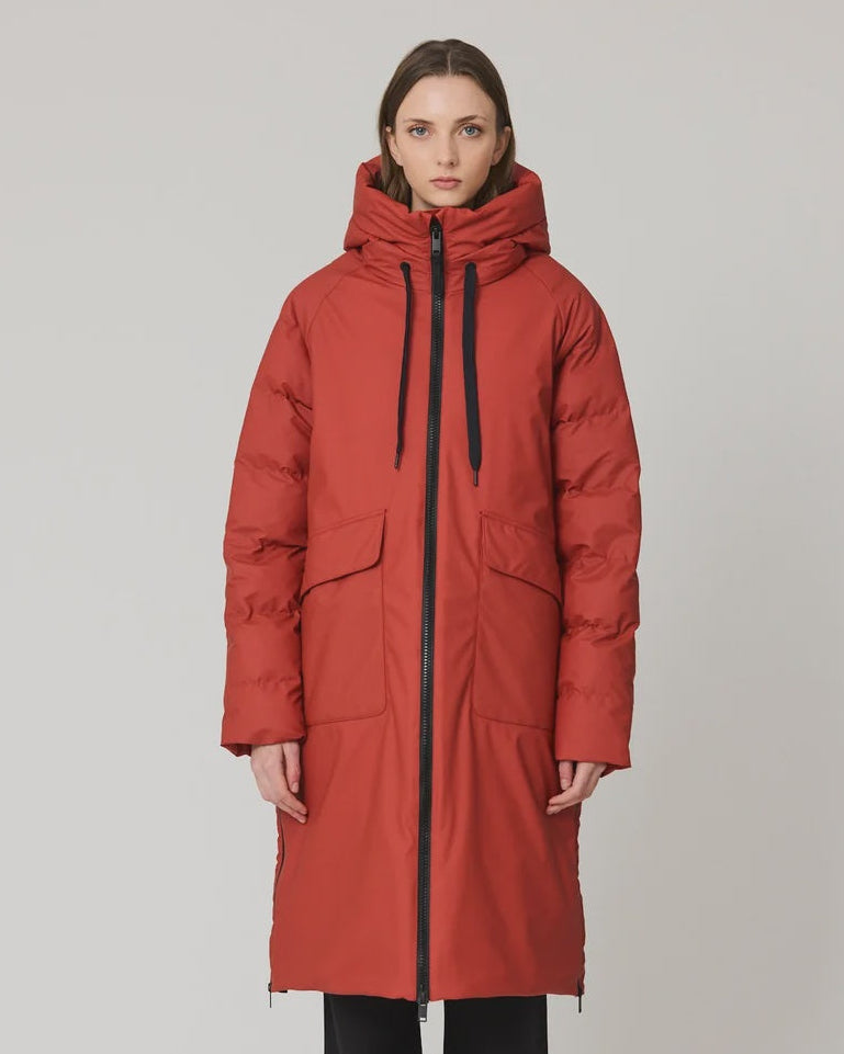 A model stands against a light gray studio background wearing a long, quilted hooded puffer coat in a muted brick red color, paired with black wide-leg trousers and dark boots.