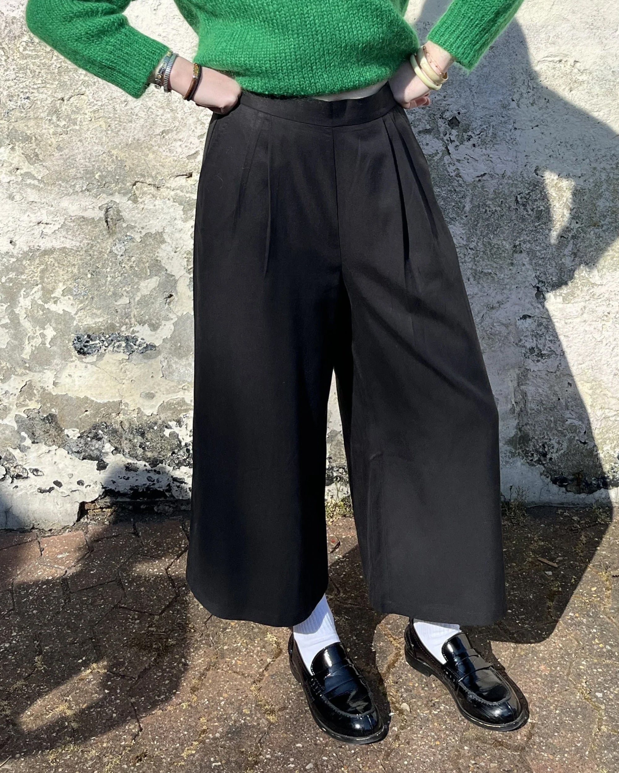 A person wearing black lyocell wide-leg trousers and a green top, standing against a textured background.