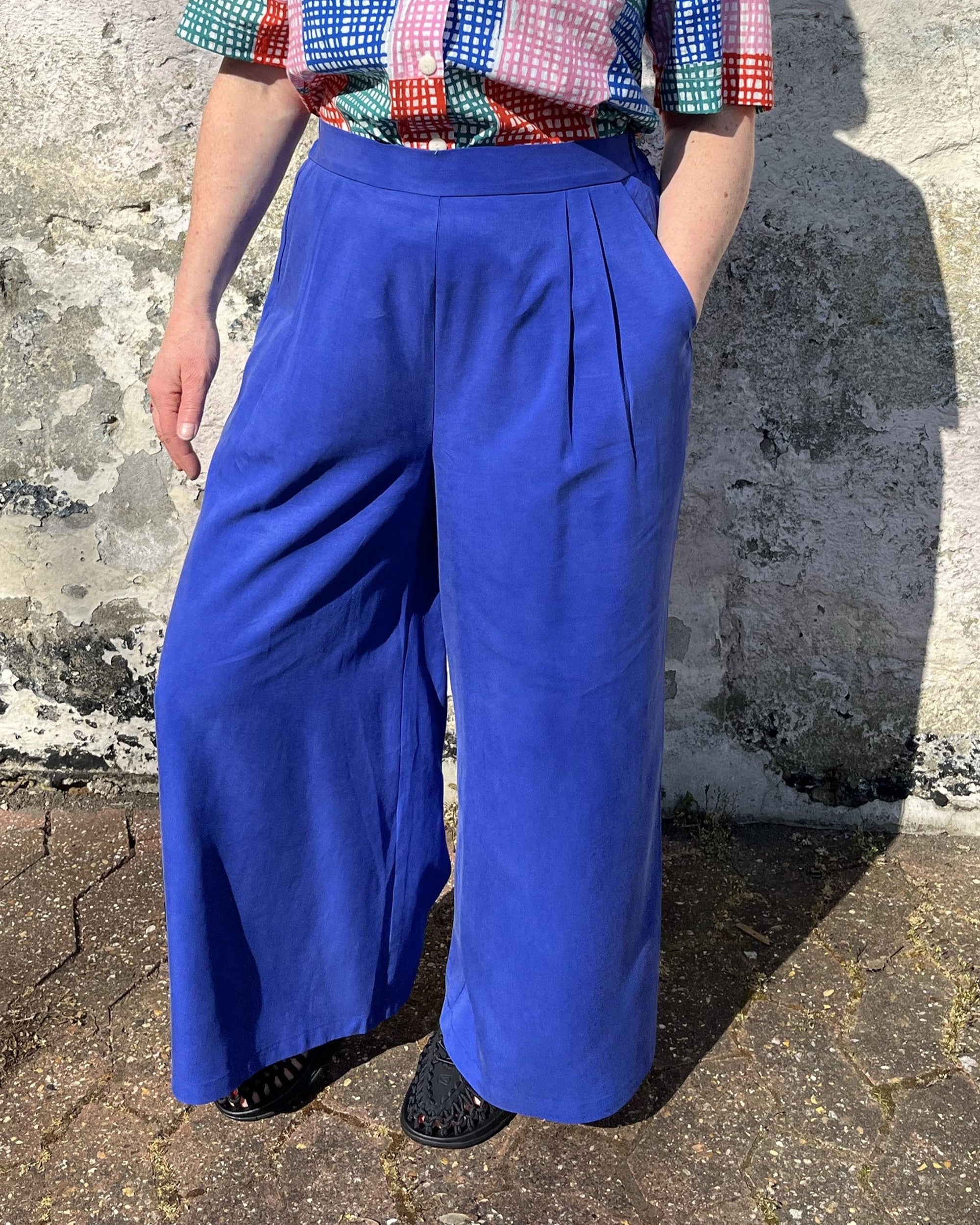 A person standing against a wall wearing cobalt lyocell wide-leg trousers with a flat front waistband, elasticated back, and pleats at the front.