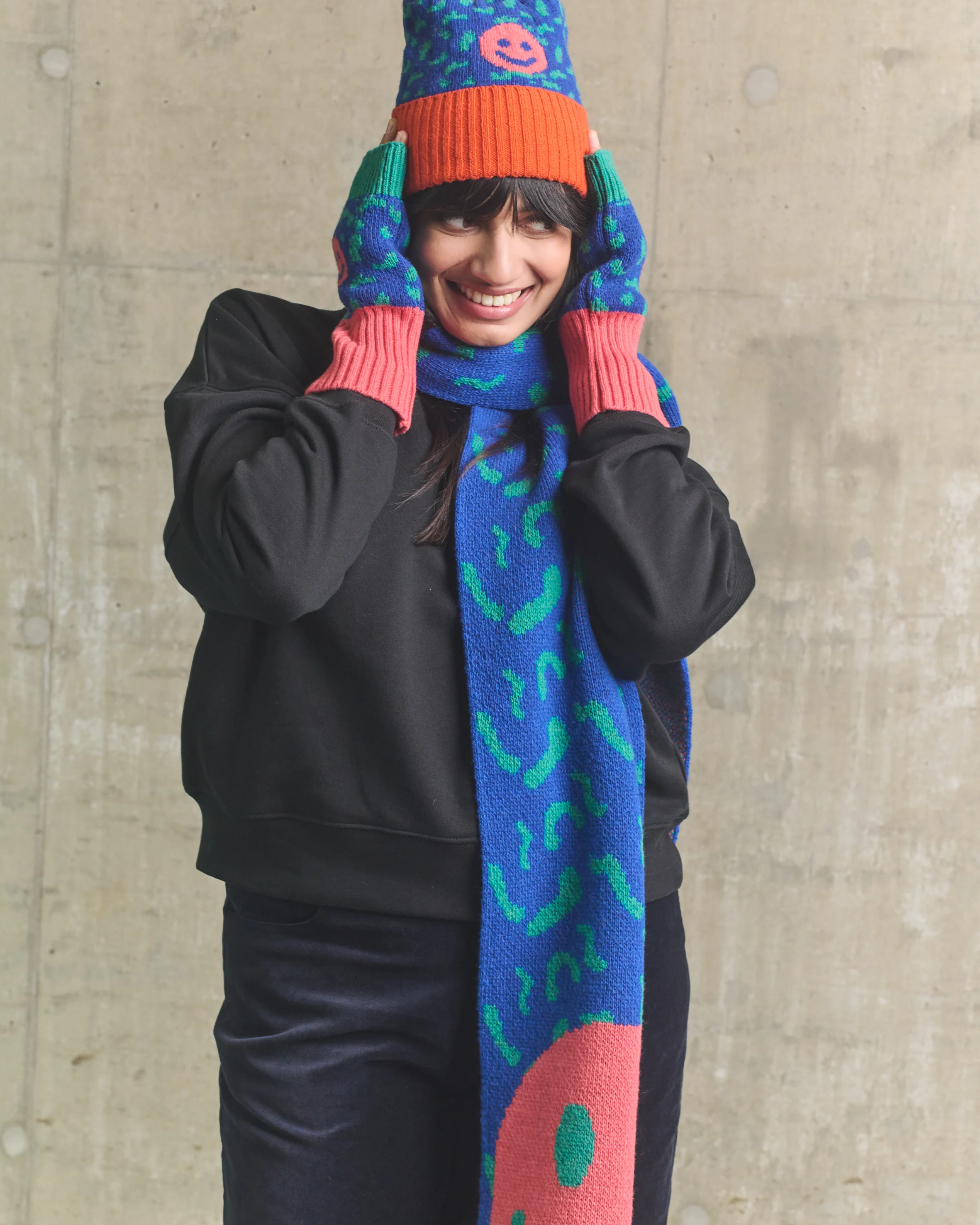 Person wearing a colorful knitted hat and scarf against a concrete wall.