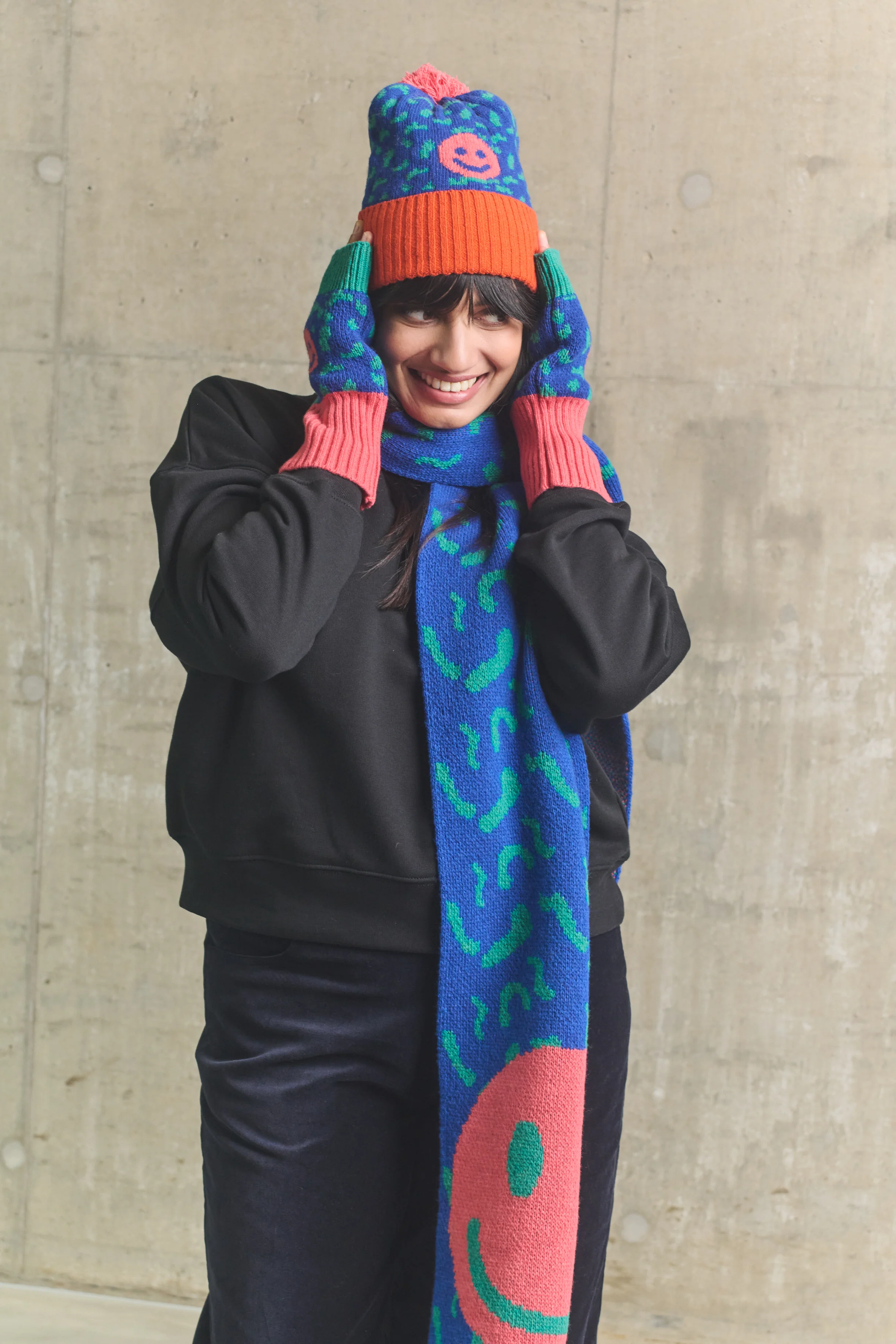 Person wearing a colorful knitted hat and scarf against a concrete wall.