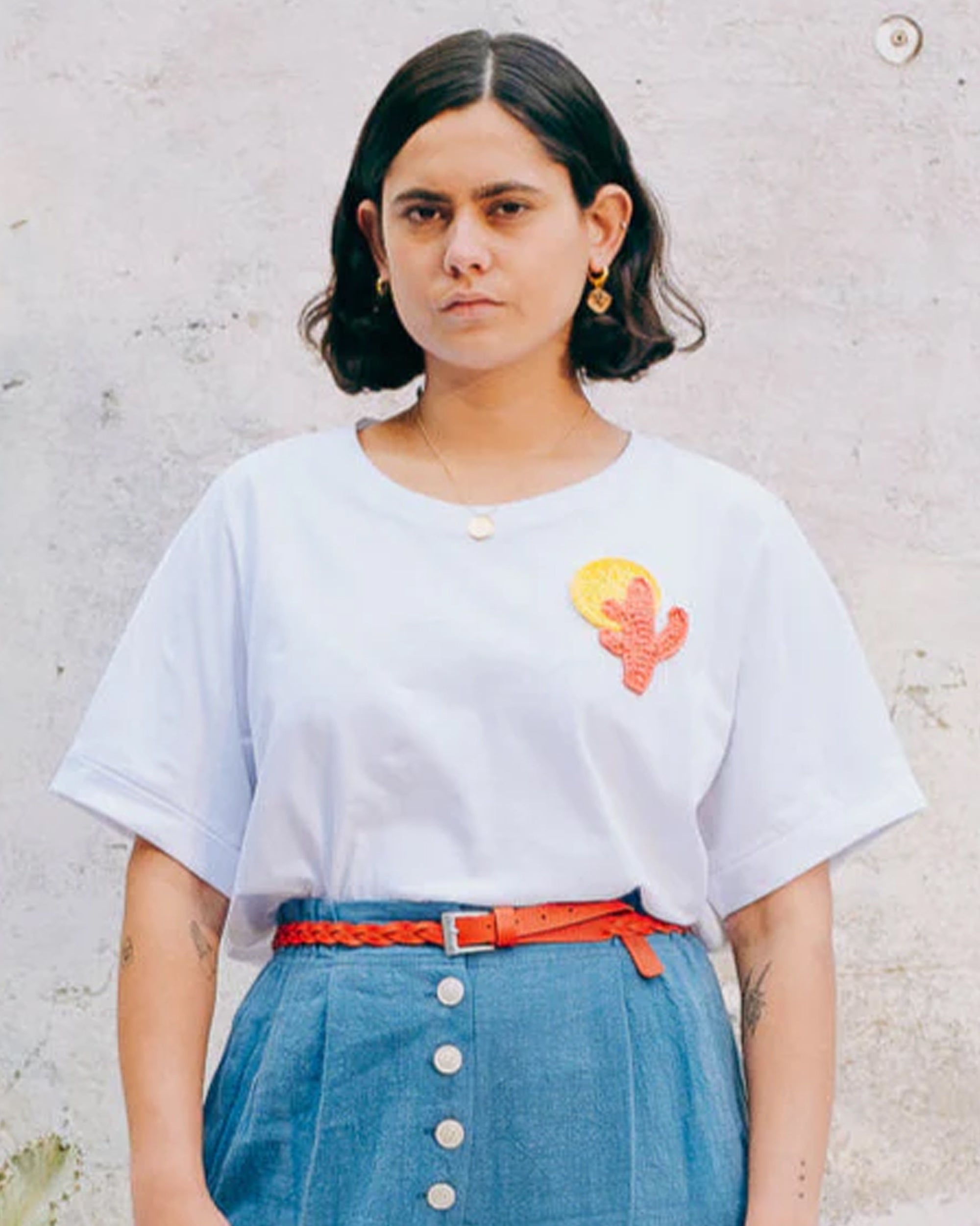 A person wearing a white crew neck t-shirt with a hand-crocheted red and yellow cactus and sun motif, paired with a blue skirt and a red belt.