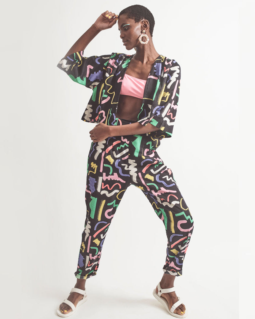 A pair of black holiday trousers with a multicolored scribble print, featuring a high waist, tapered leg, and side pockets.