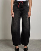A woman stands in a studio wearing a white sleeveless top and wide-leg black jeans with a red drawstring at the waist, paired with black pointed shoes.