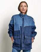 A woman stands in a studio wearing a blue patchwork denim jacket and matching blue denim cropped pants, paired with brown leather shoes, against a light gray wall and concrete floor.