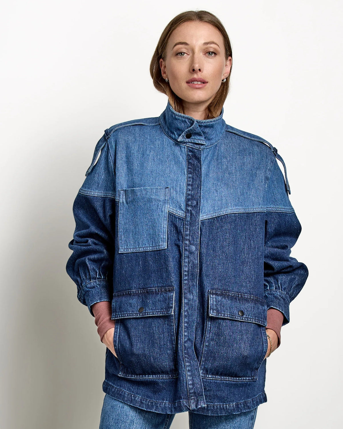 A woman stands in a studio wearing a blue patchwork denim jacket and matching blue denim cropped pants, paired with brown leather shoes, against a light gray wall and concrete floor.