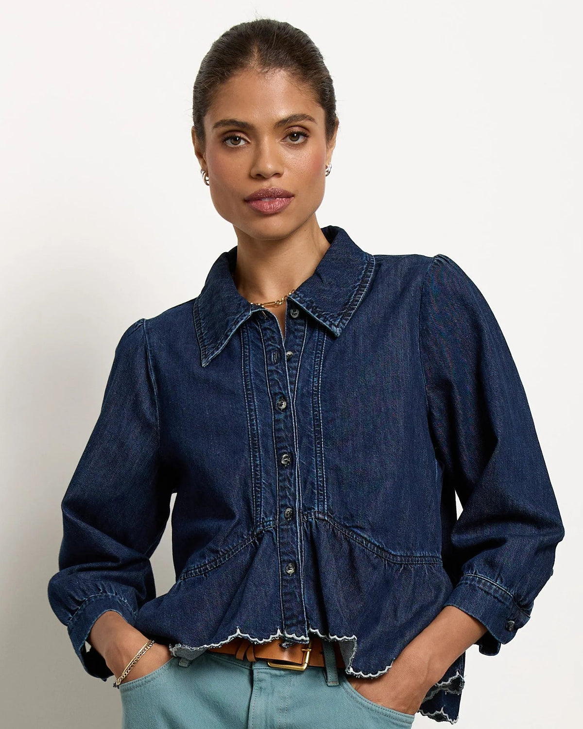 A model stands in a studio wearing a dark blue denim button-up shirt and high-waisted, wide-leg teal pants with brown leather shoes.