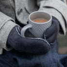 Person wearing gloves holding a white mug with a warm beverage, wearing a coat.