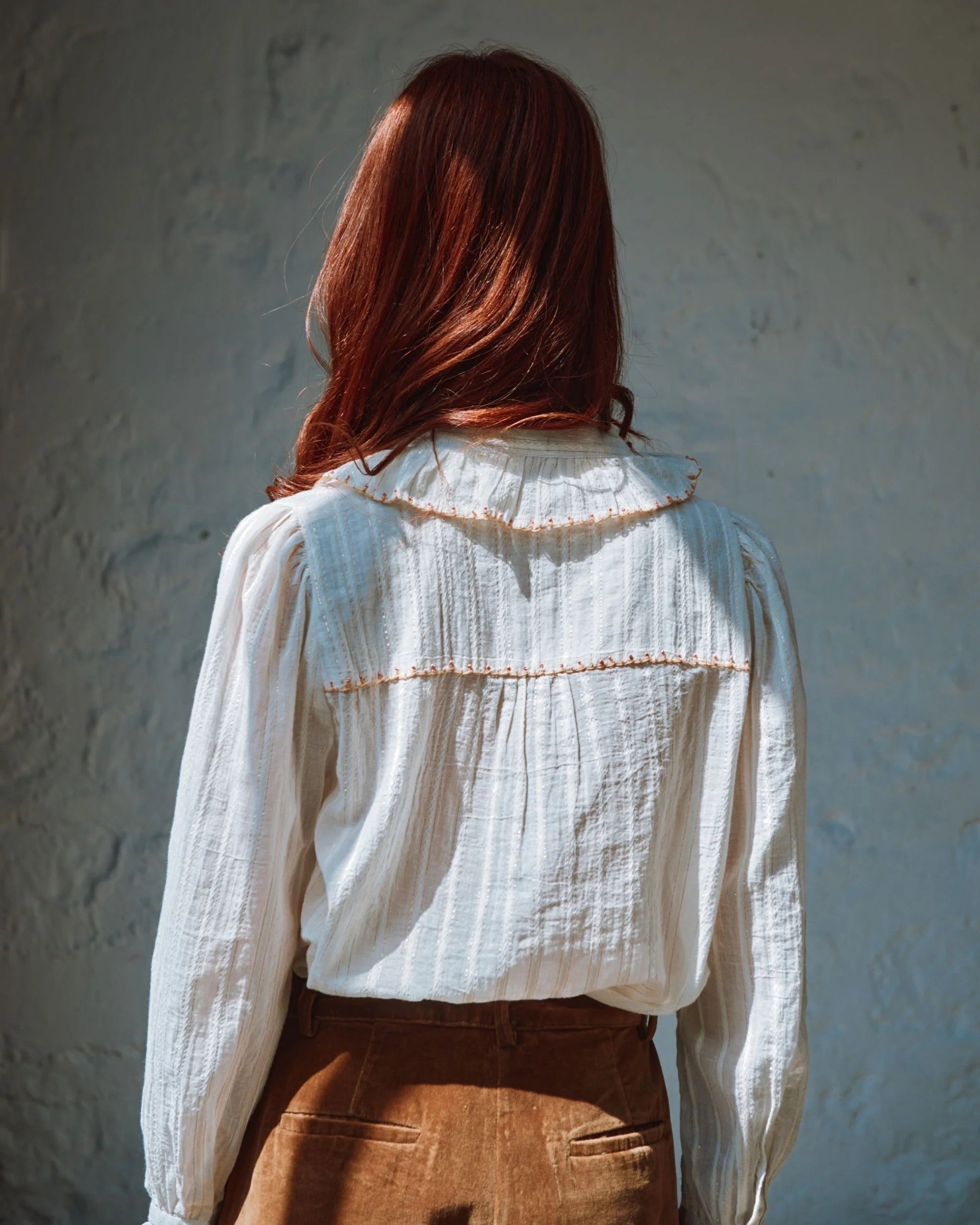 Person wearing a white blouse with brown accents against a textured wall.