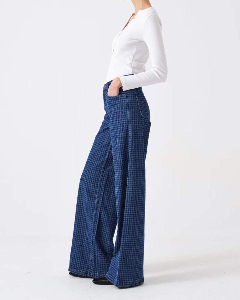 A woman stands in profile wearing a white long-sleeve top and high-waisted, wide-leg blue plaid trousers against a plain white studio background.