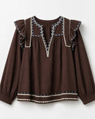 A dark brown long-sleeve blouse with ruffled shoulders and decorative embroidered trim in cream, blue, and white, shown against a light gray background.