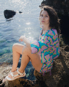 Woman in colorful patterned dress sitting on rocks by the water.