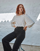 Woman wearing a polka dot blouse and black pants against a white wall.