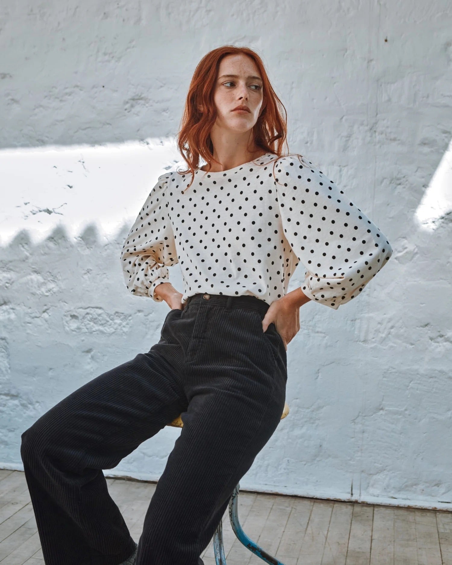 Woman wearing a polka dot blouse and black pants against a white wall.