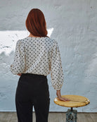 Person wearing a polka dot blouse and black pants standing next to a wooden stool against a textured white wall.