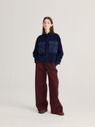 A model stands against a light gray studio background wearing a dark navy patchwork button-up shirt with wide-leg burgundy pants and brown shoes.