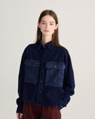 A model wearing a dark indigo denim jacket with contrasting medium-blue patch pockets on the chest, paired with burgundy pants, against a plain light gray studio background.