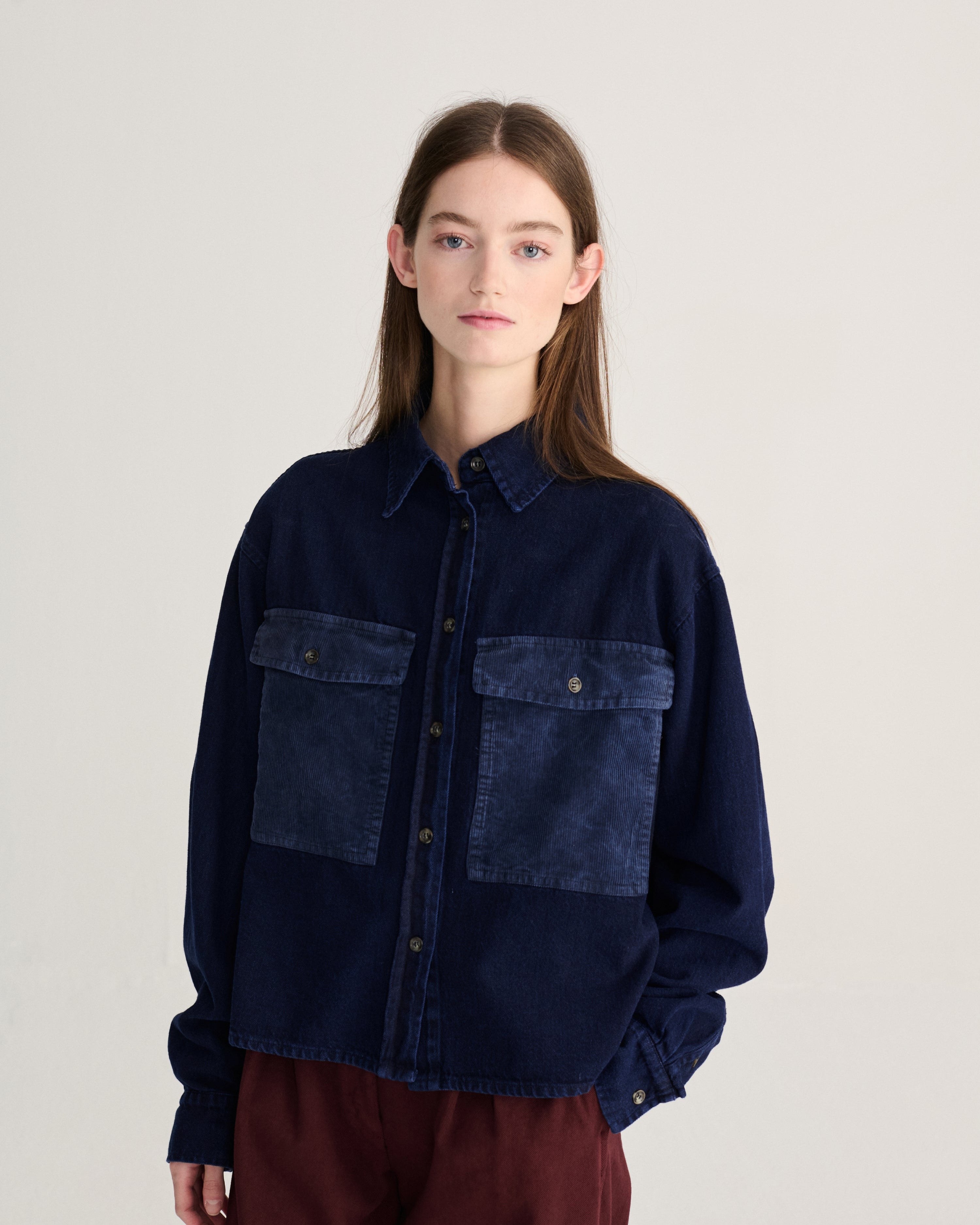 A model wearing a dark indigo denim jacket with contrasting medium-blue patch pockets on the chest, paired with burgundy pants, against a plain light gray studio background.