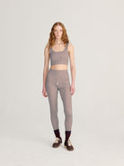 A model stands against a light gray studio background wearing a matching two-piece set with vertical stripes in dark brown, beige, and white. The set includes a sleeveless crop top and high-waisted leggings, paired with dark socks and brown sneakers.