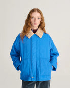 A model with long red hair wearing a bright blue quilted jacket with a beige sherpa collar, standing against a light gray studio background.
