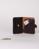 A dark brown leather wallet shown open, displaying its interior card slots, a cardholder pocket, and a snap-button closure. A small set of coins is placed on the surface to the left for scale, against a clean, light gray background.