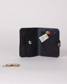 A small black textured leather wallet shown open on a light gray studio background, with a card partially inserted and a few coins placed beside it.