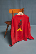 A red crewneck sweatshirt hanging on a wooden hanger from a wooden chair, showing a graphic of a figure with black hair and a green hat on the back, set against a gray studio background.