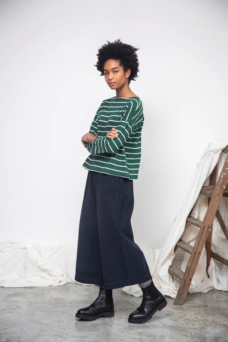 Person wearing a green striped sweater and dark blue skirt standing against a white wall with a wooden ladder.