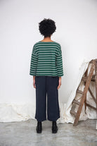Person wearing a green and white striped sweater and dark pants standing against a white wall with a wooden ladder.
