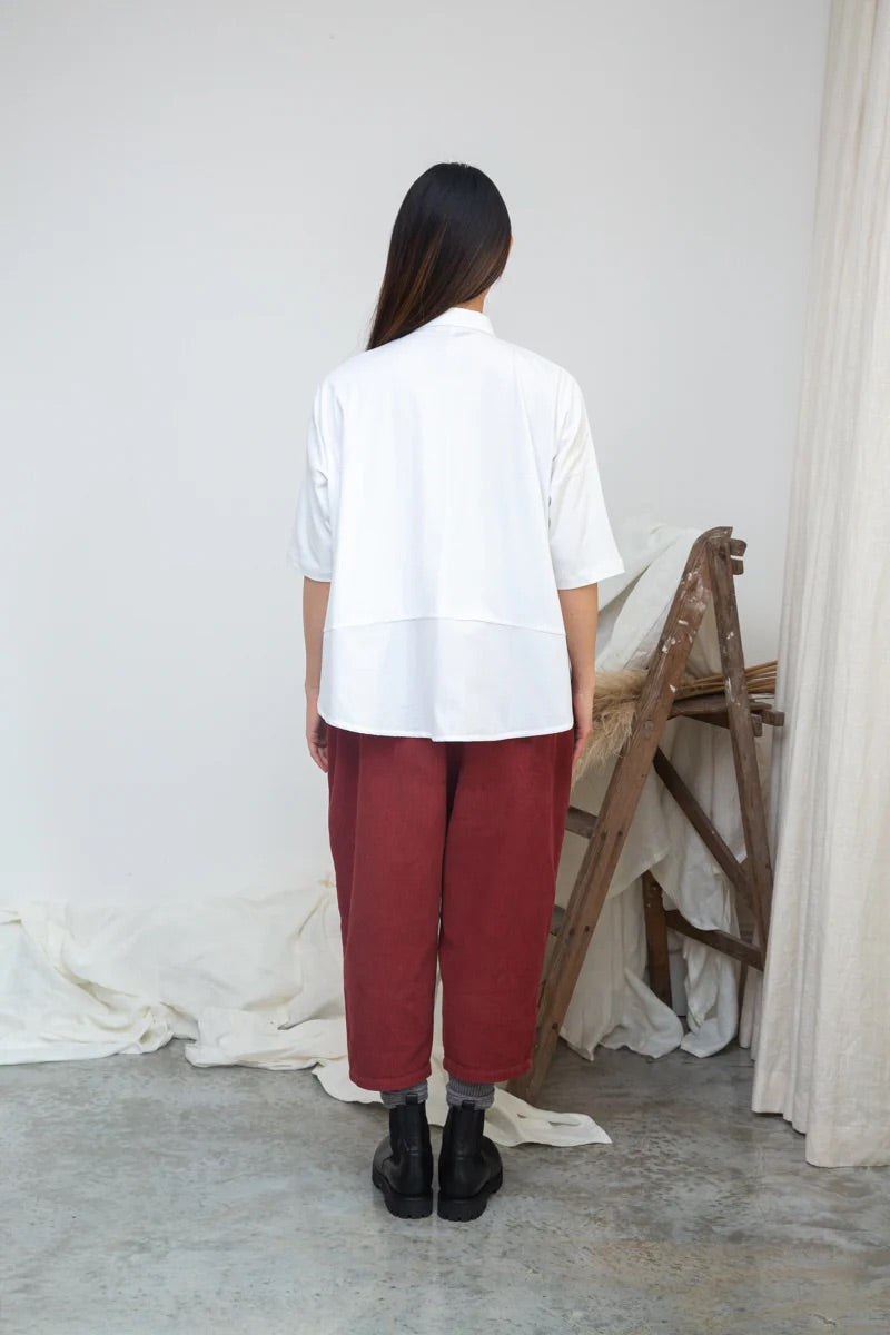 Person wearing a white shirt and red pants standing in a minimalistic room with a wooden chair and white curtains.