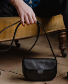 A black textured leather shoulder bag with a long strap, held by a person sitting on a mustard-yellow upholstered bench over a tiled floor.