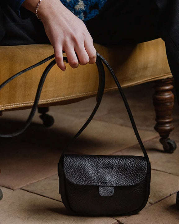 A black textured leather shoulder bag with a long strap, held by a person sitting on a mustard-yellow upholstered bench over a tiled floor.