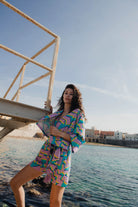 Woman in colorful outfit standing by a waterfront with buildings in the background.
