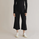 A model wearing black lounge pants with an elastic waistband and a relaxed fit. The pants are made from organic cotton and elastane, and the model is standing with one foot on a white shoe.