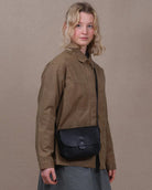 A woman stands in profile wearing a brown jacket, a gray skirt, black tights, and black shoes, with a black crossbody bag at her waist, against a plain beige studio backdrop.
