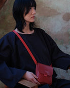 A woman with short black hair sits on a wooden chair against a textured plaster wall, wearing a black outfit and carrying a small red leather crossbody bag.