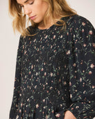 A woman wearing a dark navy floral long-sleeve blouse with a smocked bodice and peplum hem, paired with dark jeans against a plain light background.