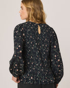 A woman shown from the back wearing a dark navy floral long-sleeve blouse with puff sleeves and a small keyhole cutout at the neckline, paired with dark jeans against a light gray studio background.