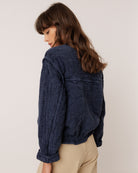 A woman with long brown hair is shown from a side-back angle wearing a dark navy crinkled textured jacket and beige trousers, photographed against a plain off-white studio background.